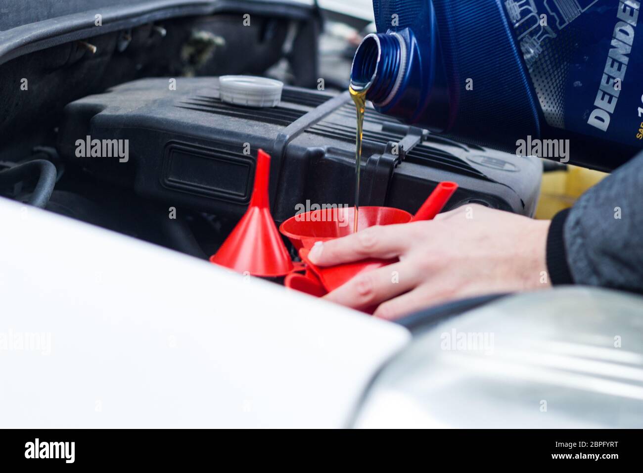 Man uses a red funnel to fill the engine oil in a car with a blue canister. Mechatronic oil level expert e-mobility Stock Photo