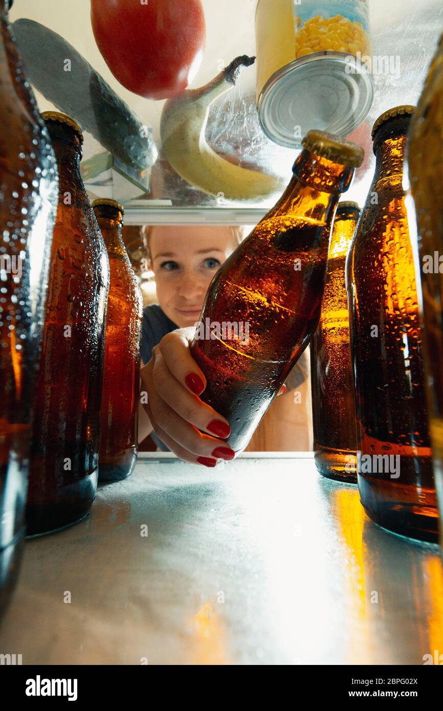 Caucasian woman takes cold refreshing beer from out the fridge, inside