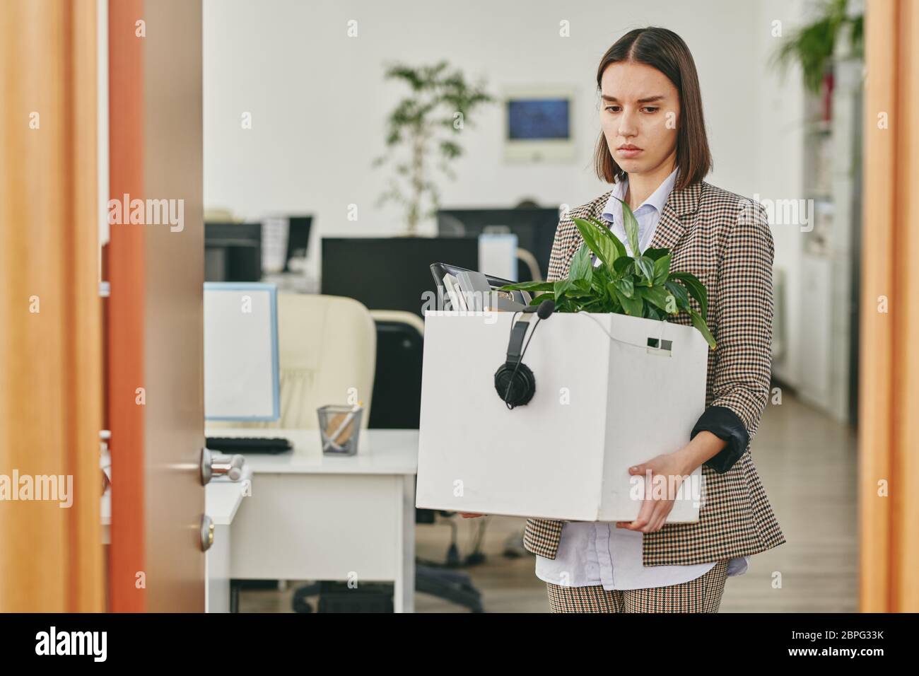 Sad young woman carrying box full of personal stuff while leaving office after retrenchment in coronavirus crisis Stock Photo