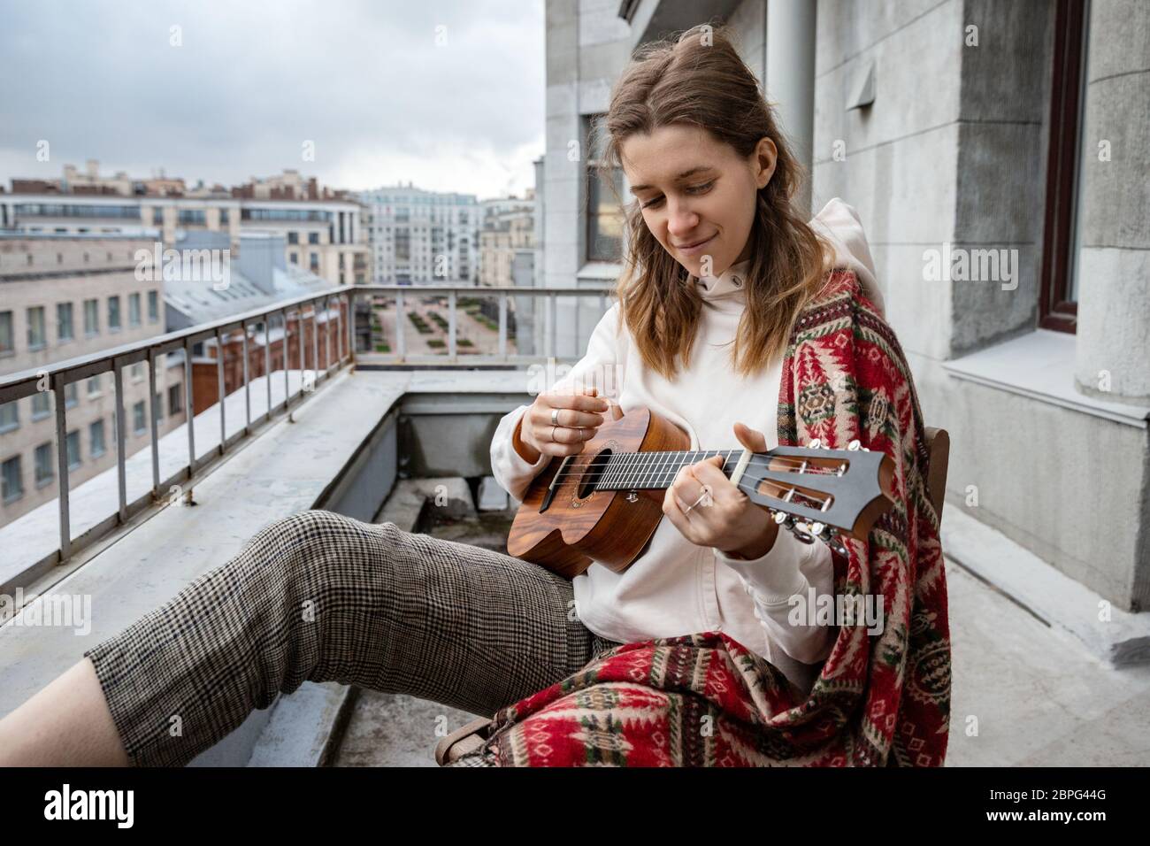 Young caucasian hipster/hippie woman in casual clothes playing on Hawaiian guitar, sings a song on a ukulele during self-isolation in the house on ter Stock Photo