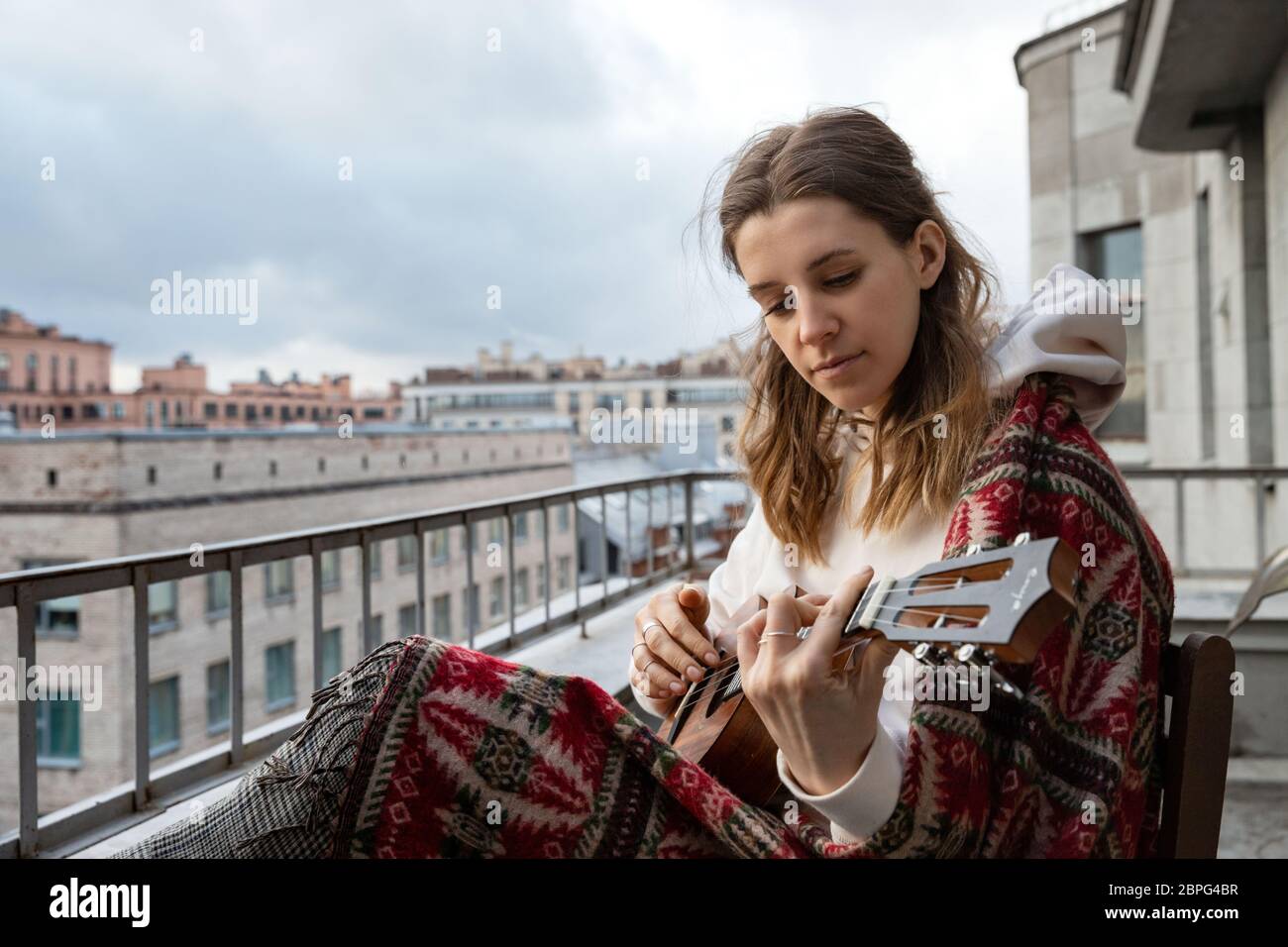 Young caucasian hipster/hippie woman in casual clothes playing Hawaiian guitar, sings a song on a ukulele during self-isolation in the house on terrac Stock Photo