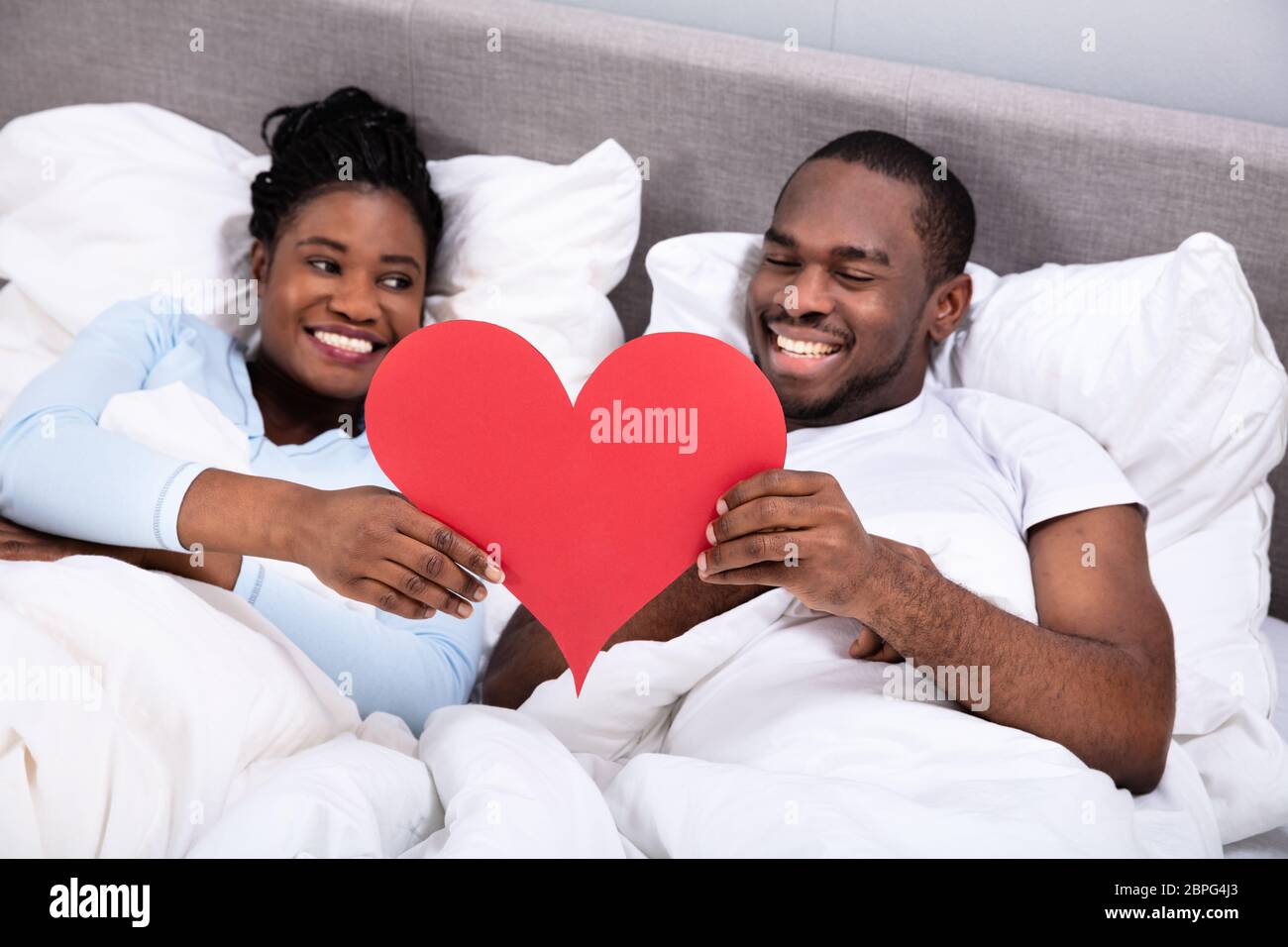 Smiling Young African Couple Lying On Bed Holding Red Heart Stock Photo ...
