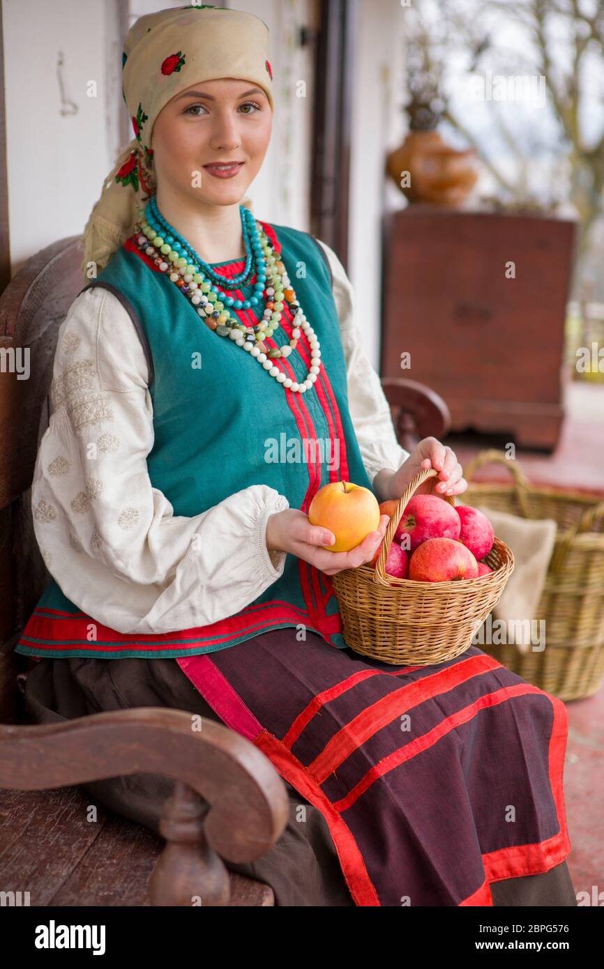 A beautiful girl in national dress is sitting. With apples in a basket. In old clothes of the 19th century. With a wreath and ribbons. Against the Stock Photo