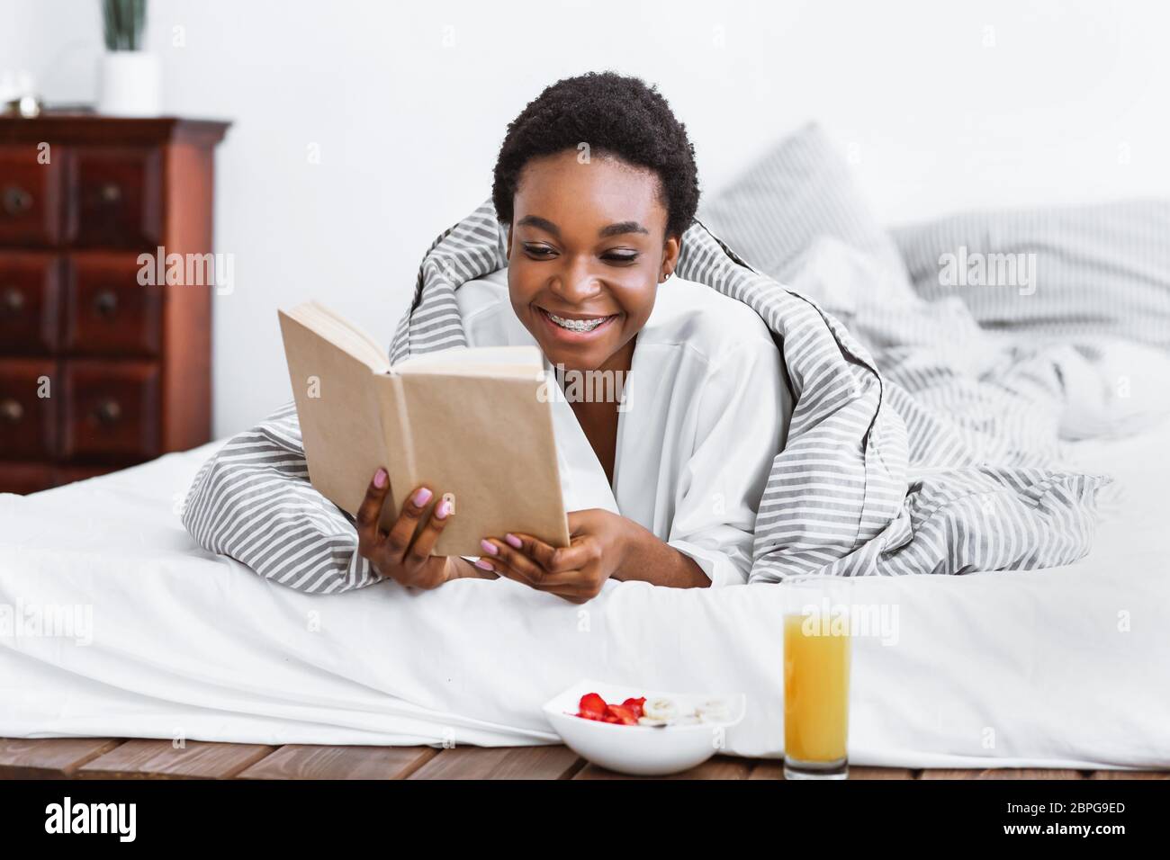 Happy african american girl reading book in bed, breakfast near Stock Photo
