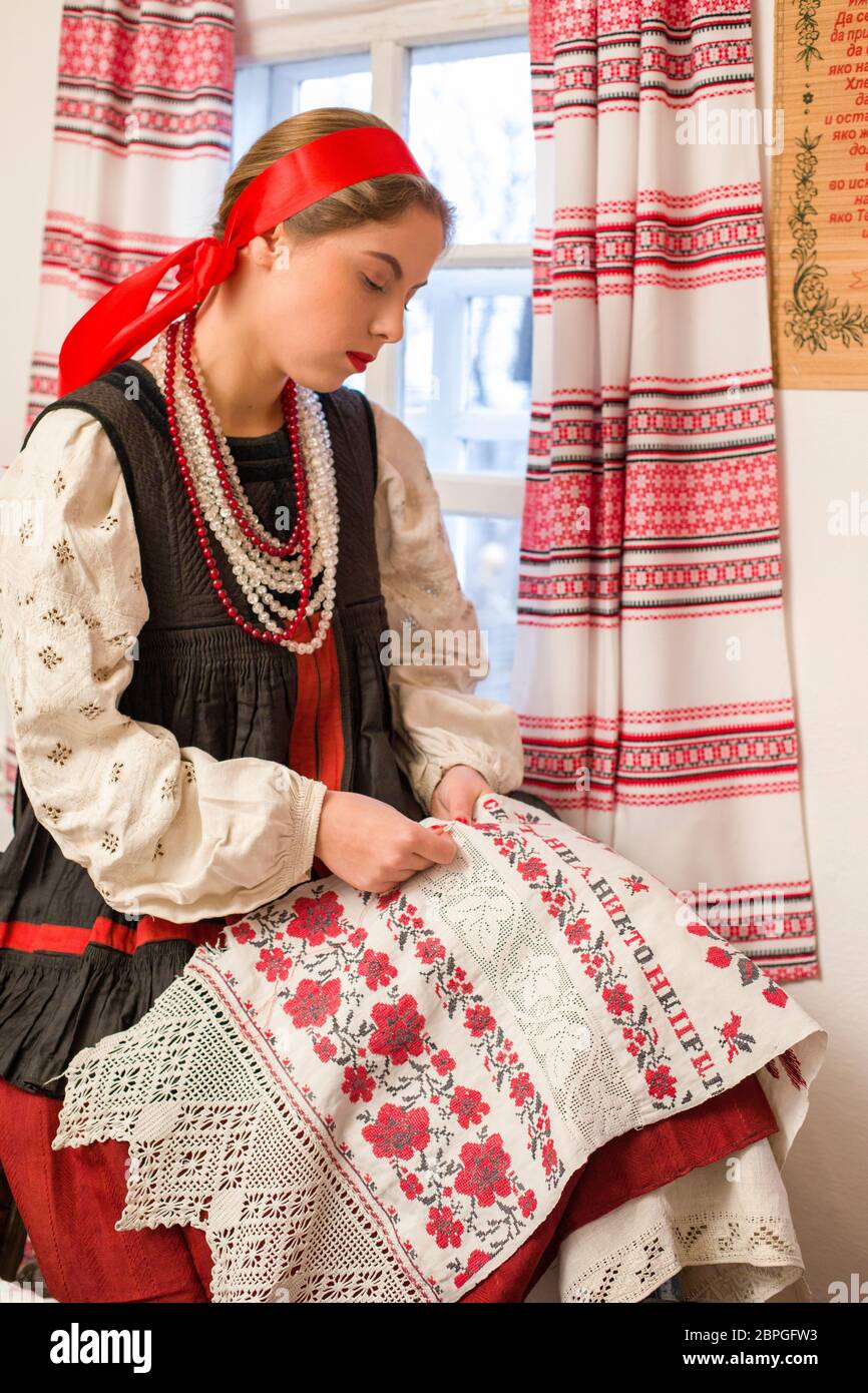 Beautiful young girl in a national costume of the 19th century. Embroiders a beautiful pattern near the window. With a wreath and ribbons. Village Stock Photo