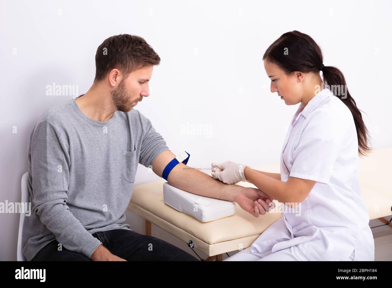 Young Female Doctor Injecting Male Patient With Syringe To Collect Blood Sample In Clinic Stock Photo
