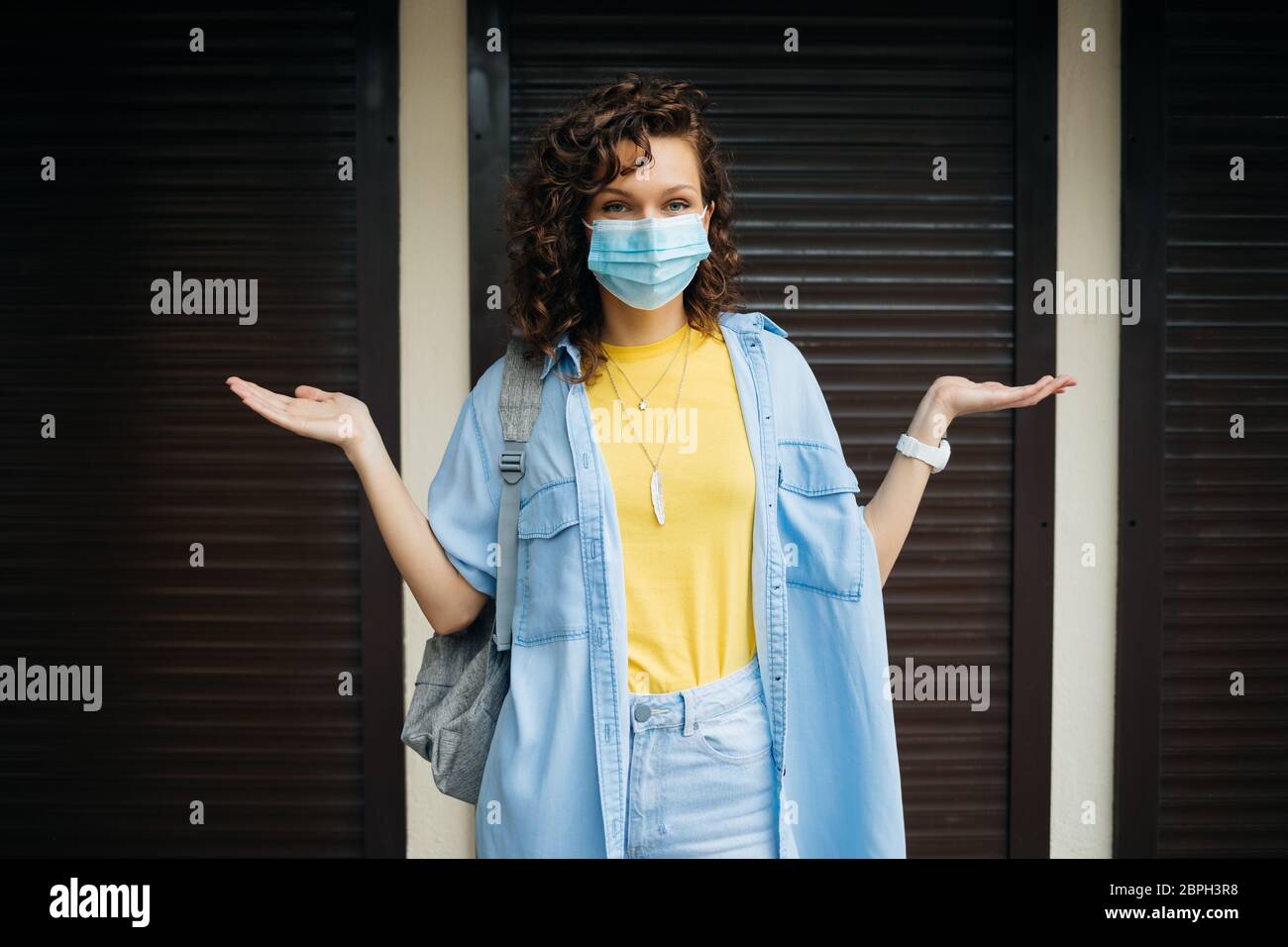 Closed stores during quarantine, young woman in protective medical mask stands on the threshold of non-working shop in confusion. Stock Photo
