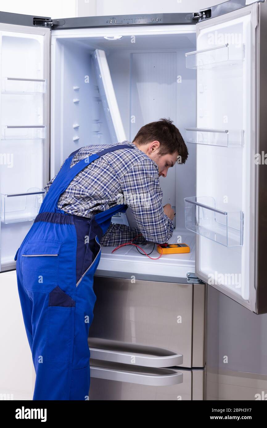 Young Serviceman Checking Fridge With Digital Multimeter Stock Photo