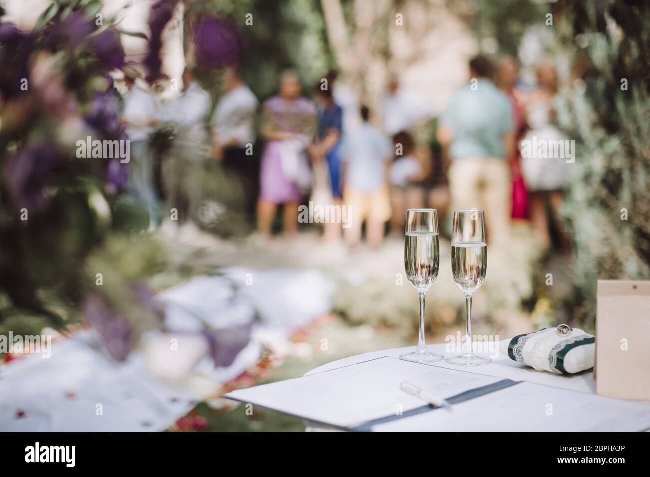 Two glasses of champagne stand on a table stand on a table. Nearby lies a pillow with wedding rings. Amid a lot of people Stock Photo