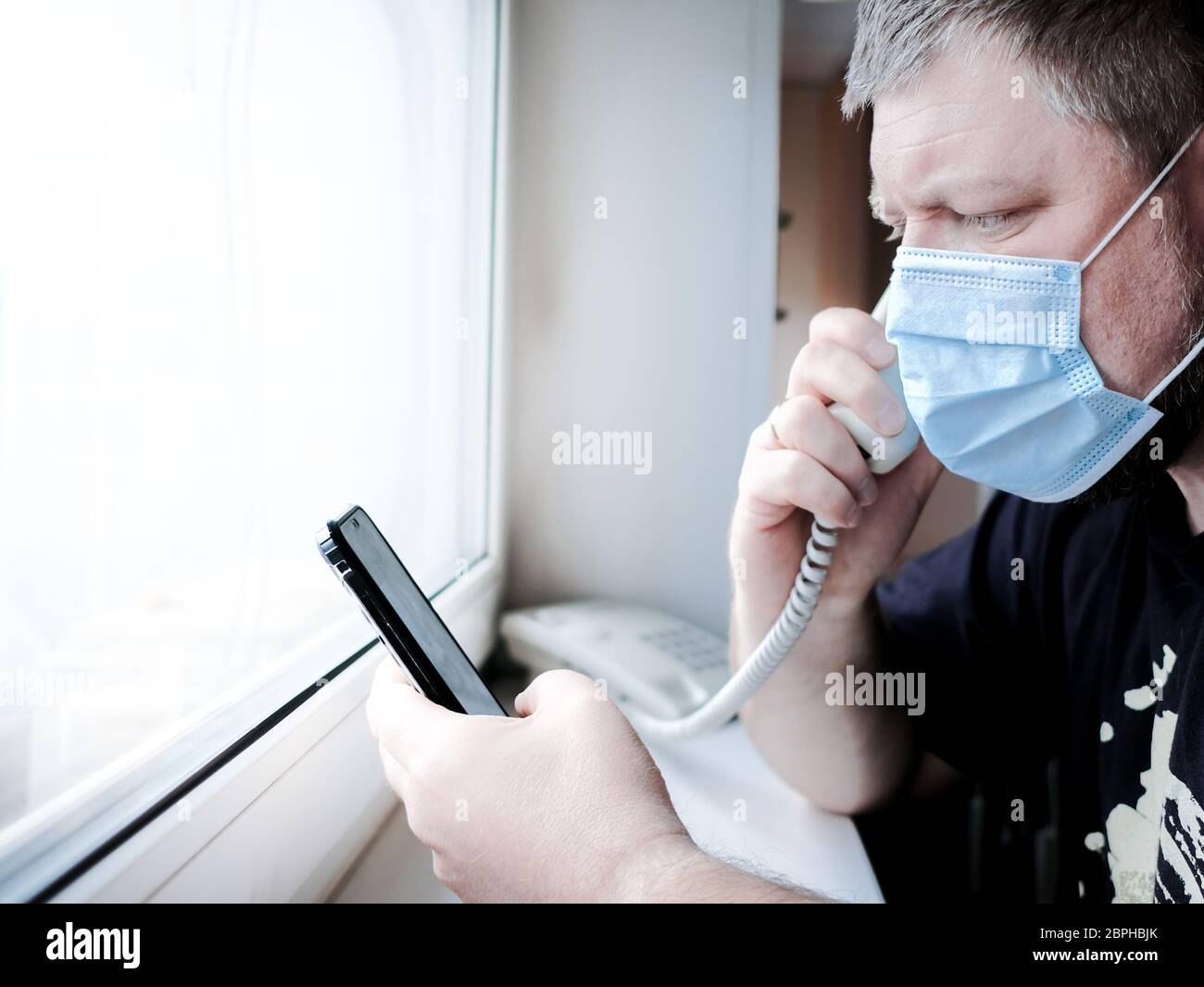 A man in a medical mask sits in front of a window and talks on a landline phone, holding a smartphone in his hand. He worries about family health duri Stock Photo
