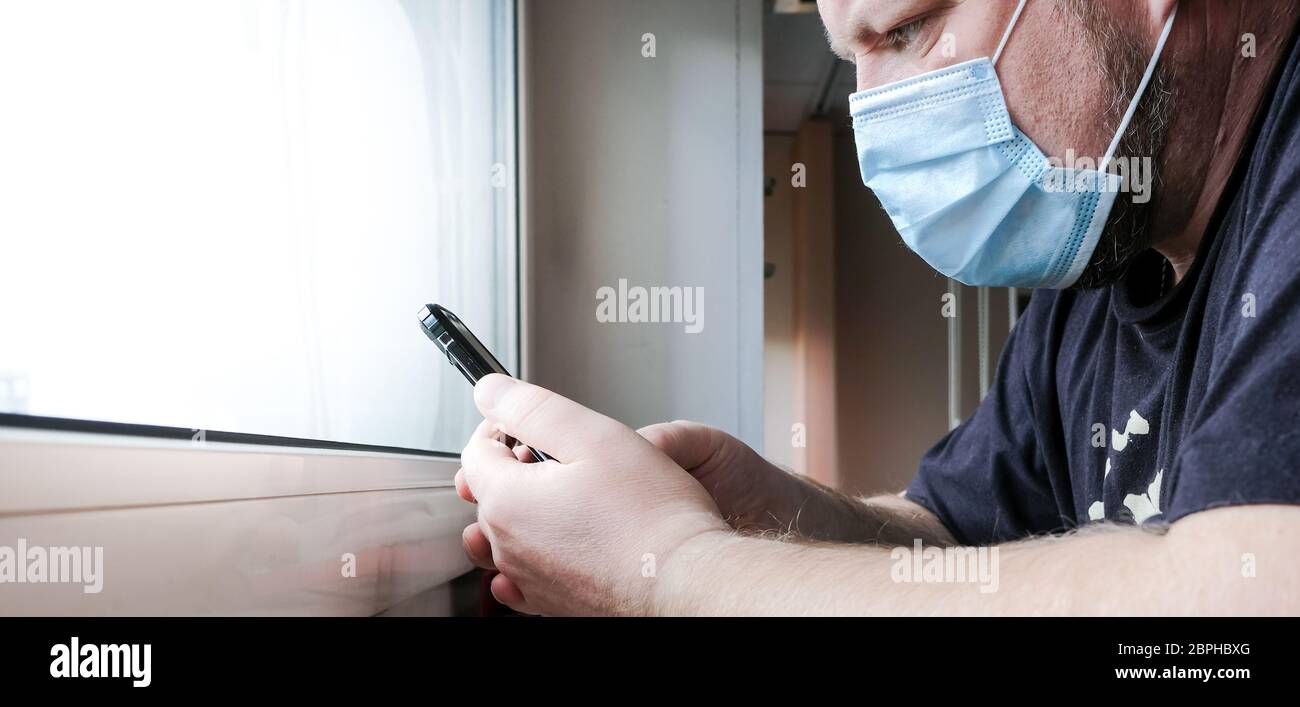 A man in a medical mask sits in front of a window and looks anxiously at a smartphone. He worries about family health during the quarantine of the cor Stock Photo