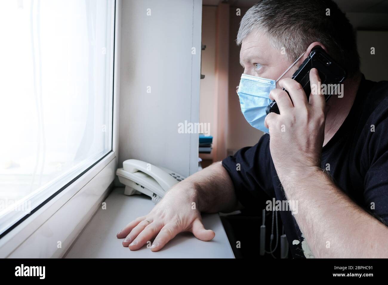 A man in a medical mask sits in front of a window and talks on a smartphone. In the eyes of man, anxiety and fear. He worries about family health duri Stock Photo