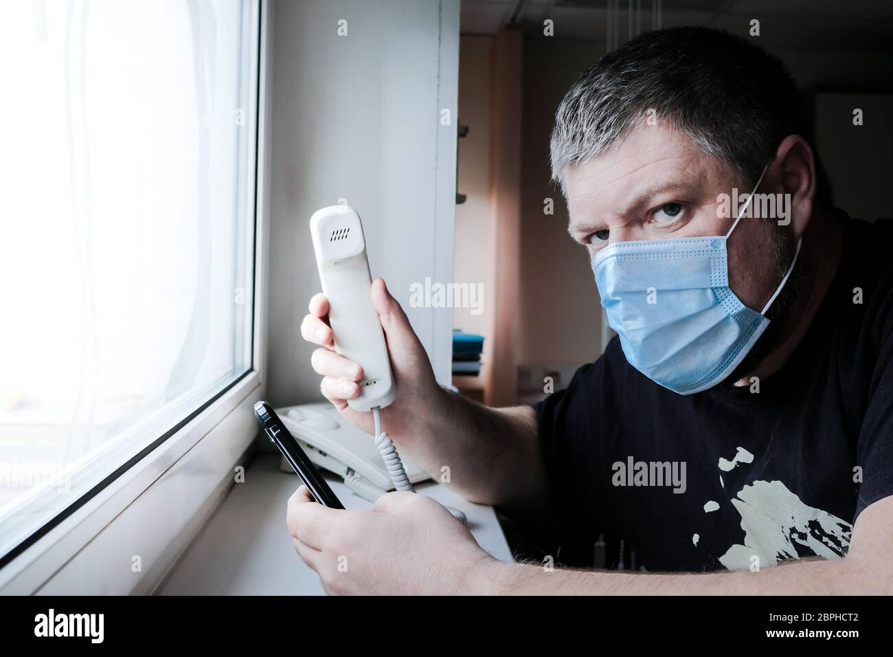A man in a medical mask sits in front of a window and holds in his hands a stationary telephone and a smartphone. He worries about family health Stock Photo