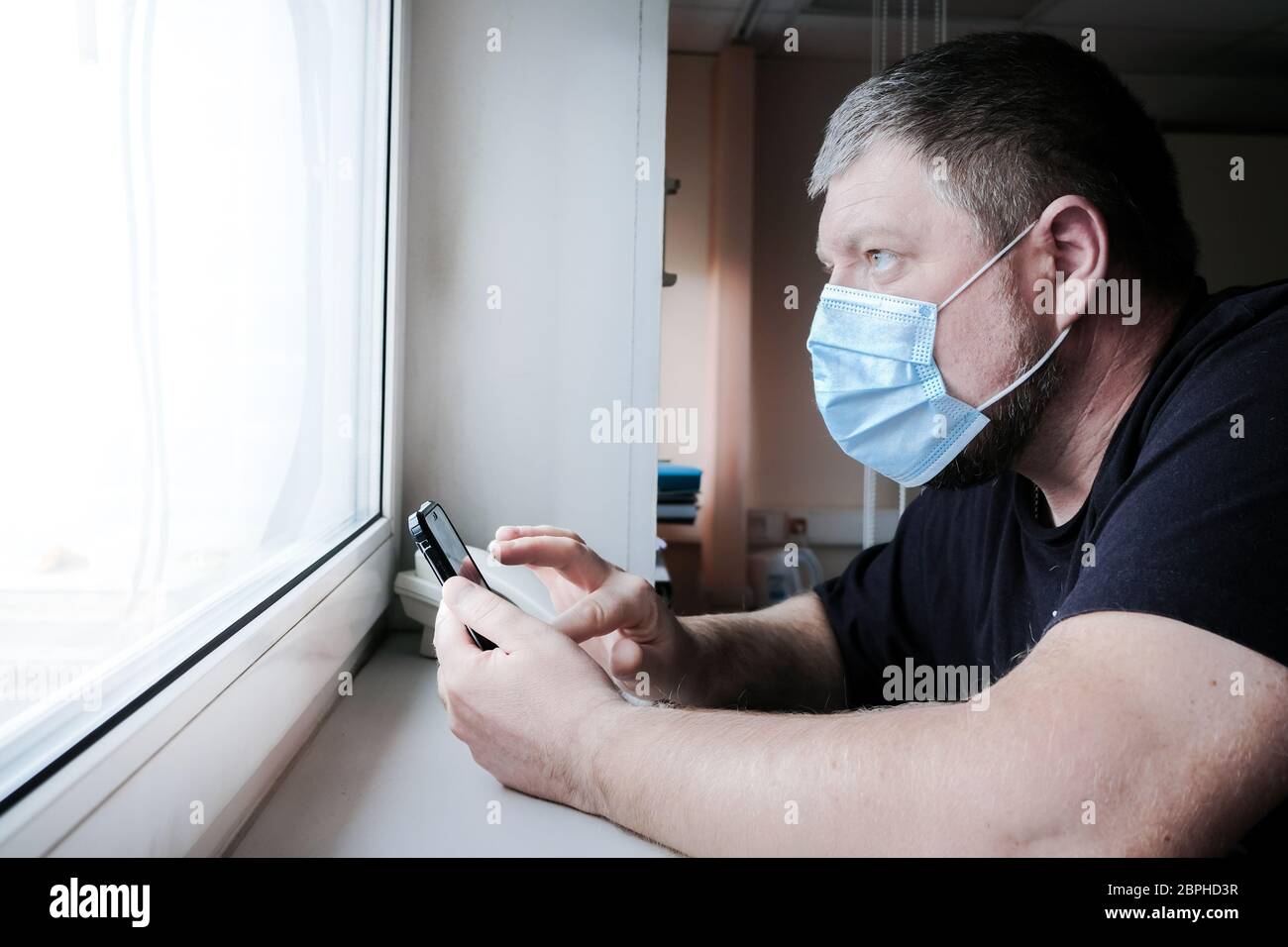 A man in a medical mask in front of the window holds a smartphone in his hands and looks anxiously out the window. He worries about family health duri Stock Photo