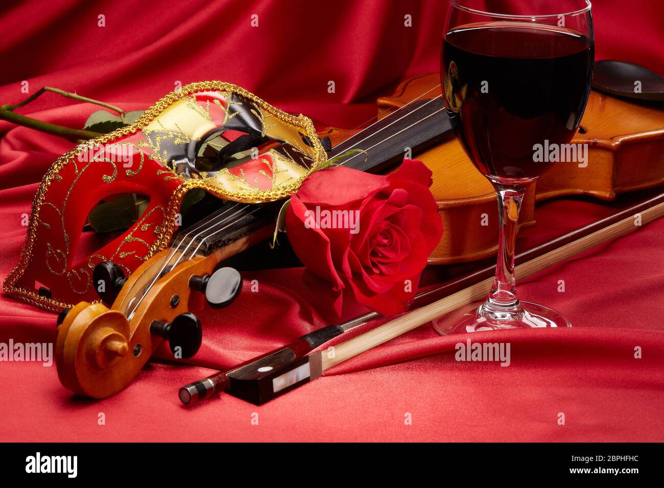 Violin (fiddle), theater mask, glass of red wine and red rose lying on the perfect red satin fabric. String instrument. Stock Photo