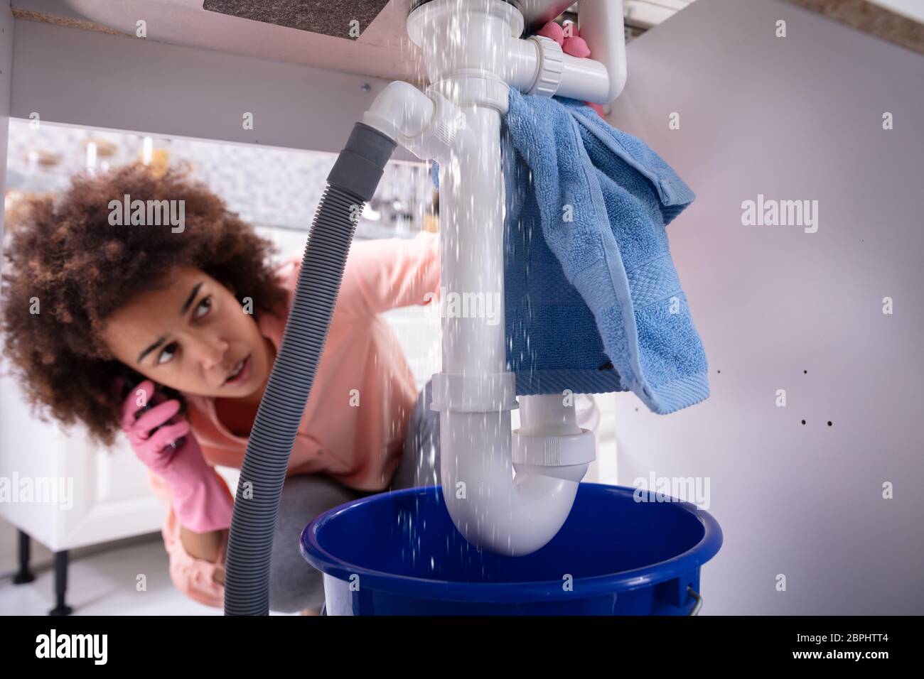 Close-up Of A Young Worried Woman Calling Plumber To Fix Sink Pipe Leakage In Kitchen Stock Photo