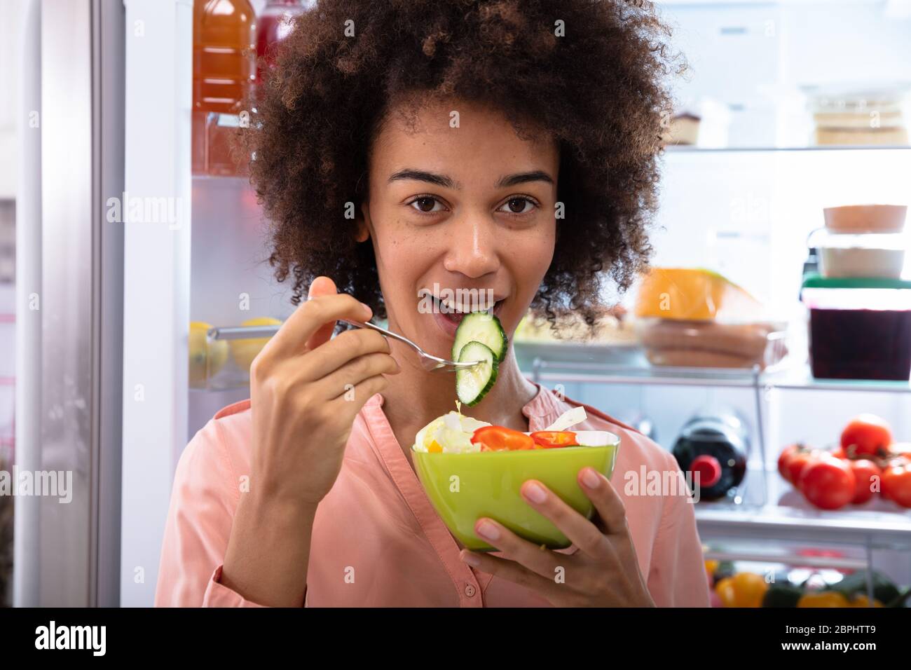 Happy Young Woman Standing In Front Of Refrigerator Eating Salad With