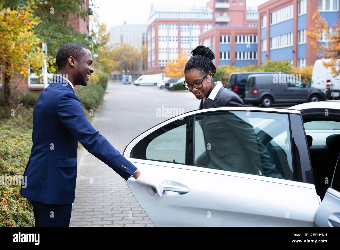 Happy African Male Valet Opening Door For Businesswoman Getting Out Of A Car On Street Stock Photo