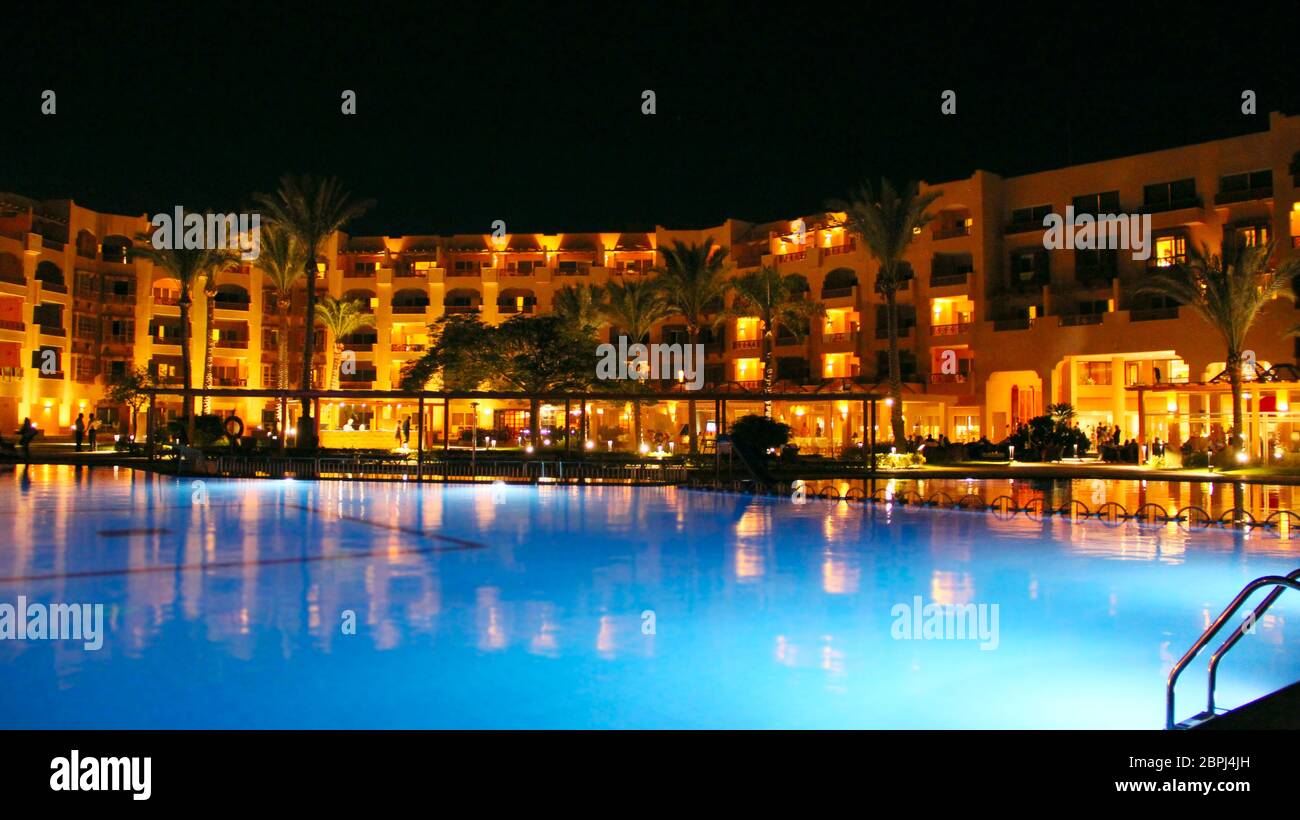 Swimming pool and evening hotel on holidays. People relax in evening near pool. Lights of evening hotel are reflected in pool water in night. Bright l Stock Photo