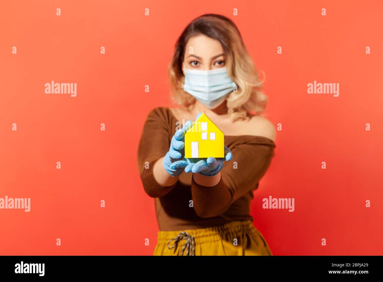 Woman in protective mask and gloves showing paper house on her hand, recommendation to stay home, adhere quarantine while contagious disease spread, c Stock Photo