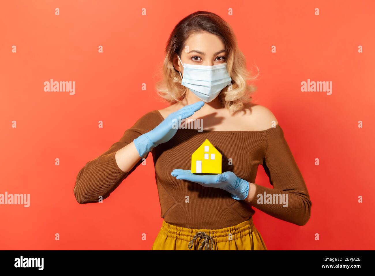 Blond woman in protective mask and gloves holding paper house as symbol of safety and care, recommendation adhere quarantine while contagious disease Stock Photo