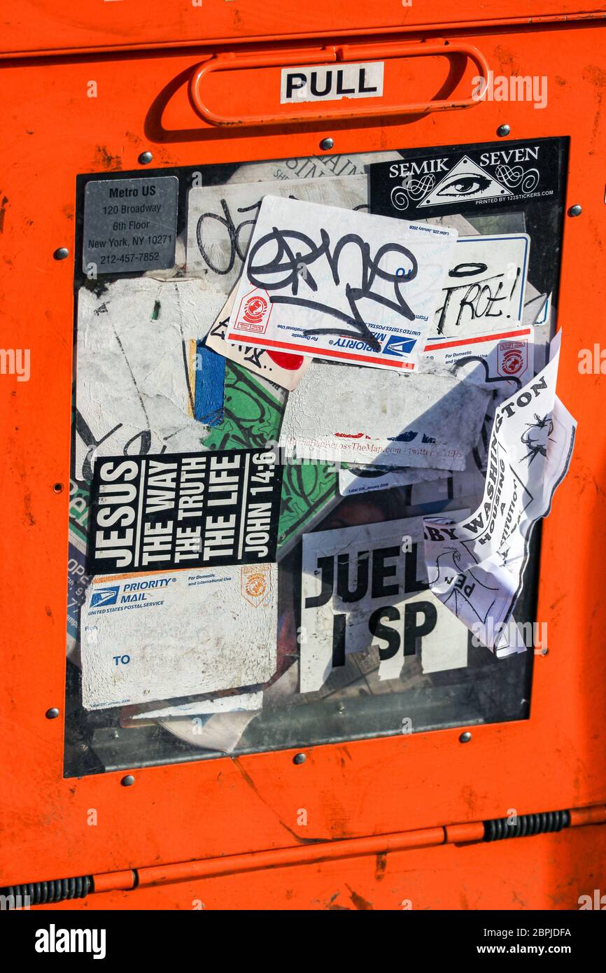Orange free newspaper dispenser covered in stickers in New York City, United States of America Stock Photo