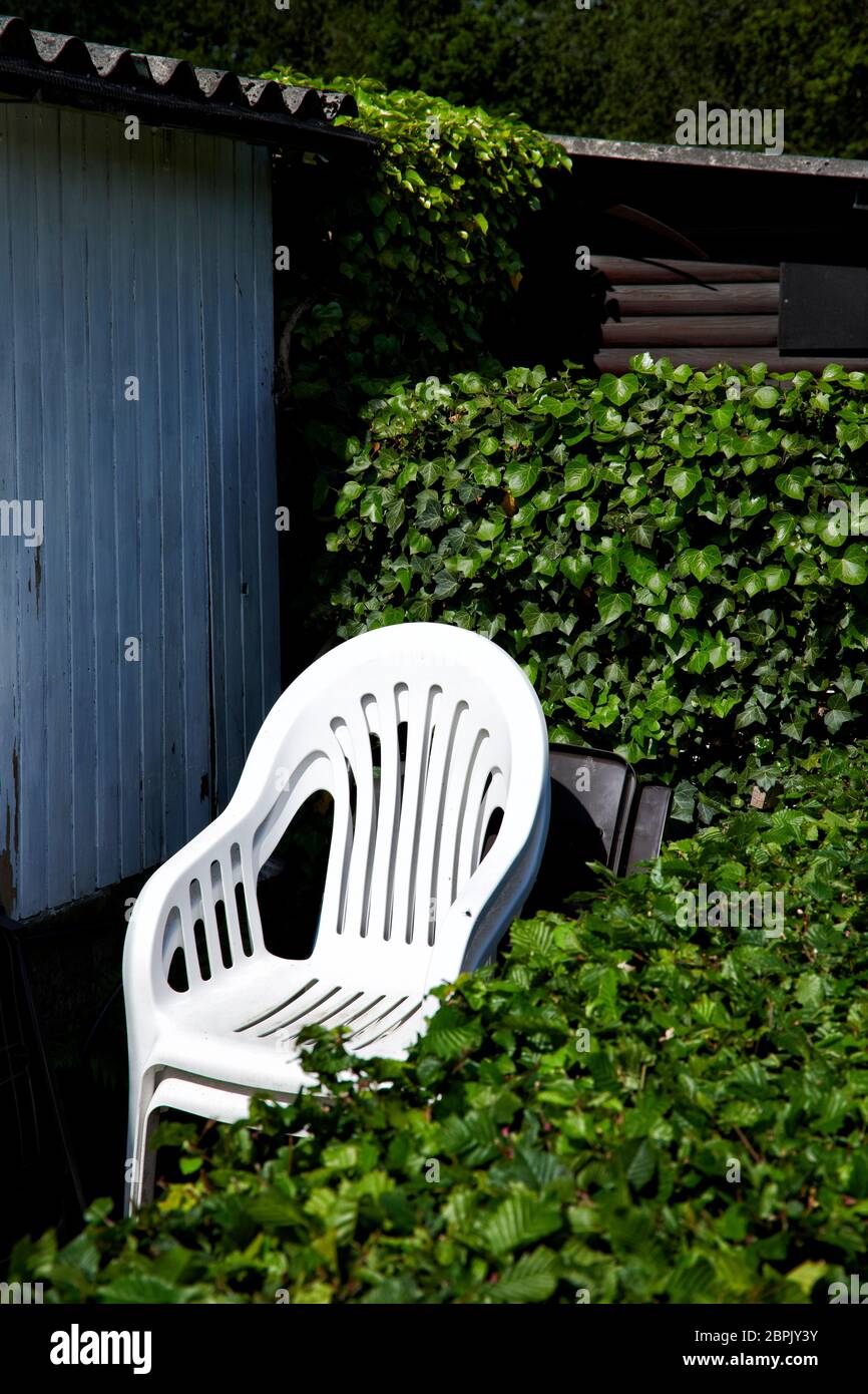 Ein Stapel weisser Kunststoffstuehle steht an der Seite einer Gartenlaube und ist umringt von einer Hecke. Stock Photo