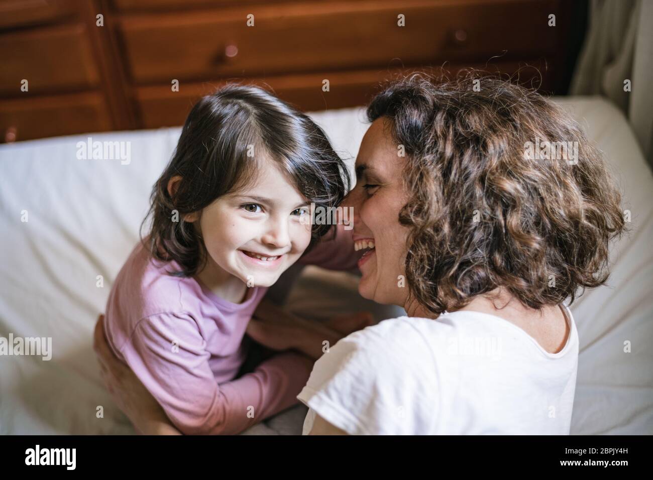 Young mother and daughter playing in the bedroom. Mom whispers into the child's ear as they both laugh Stock Photo