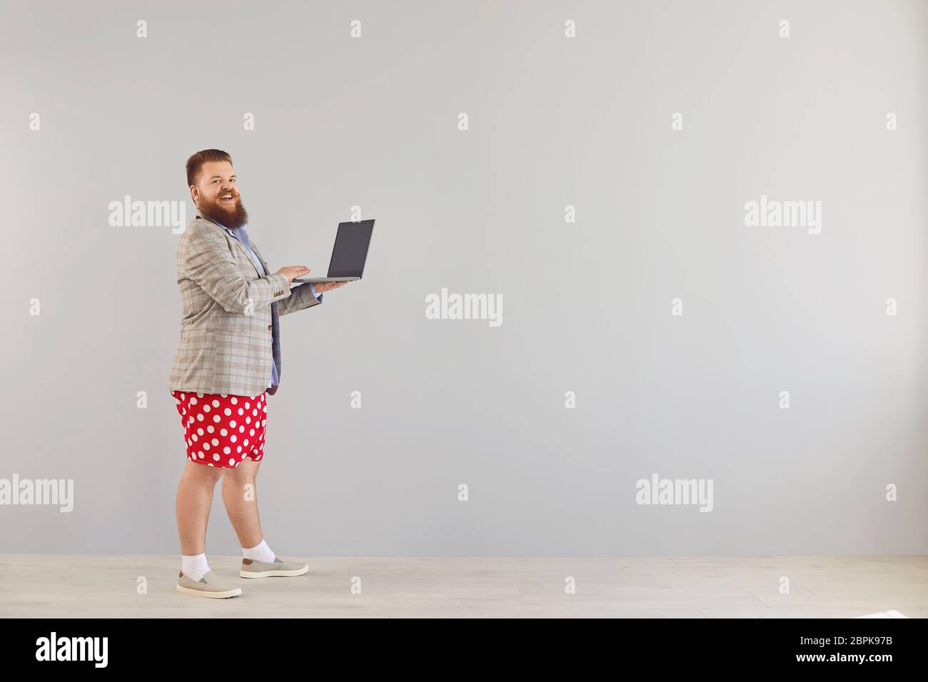 Funny fat man in a blue shirt and red shorts standing working online using a laptop on a gray background. Stock Photo