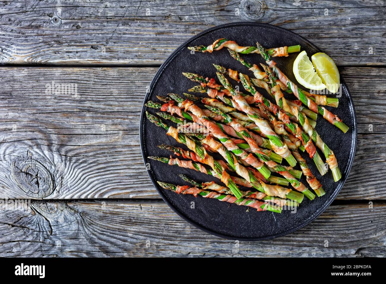 Bacon wrapped asparagus easy side dish grilled with garlic and sea salt served on a black plate lemon on an old wooden background, top view, flat lay, Stock Photo