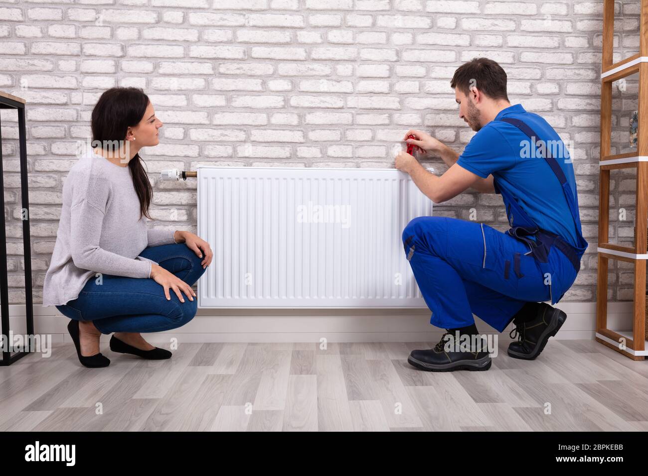 Beautiful Woman Looking At Young Male Plumber In Uniform Installing Radiator With Screwdriver Stock Photo