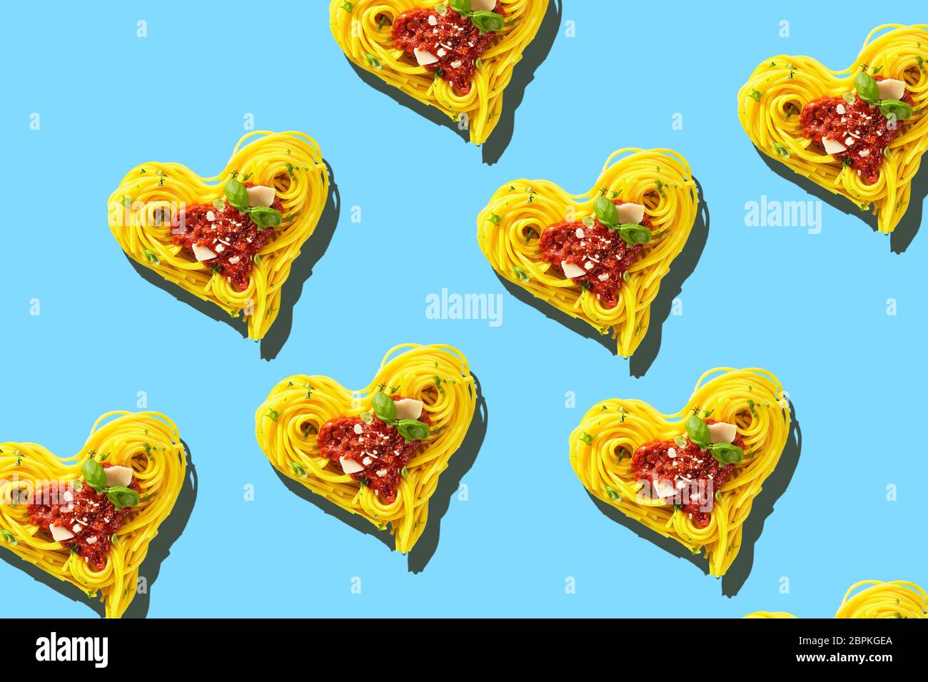 Decorative hearts of pasta of cooked yellow spaghetti with tomatoes, basil and parmesan cheese toping, viewed from above in full frame on light blue b Stock Photo