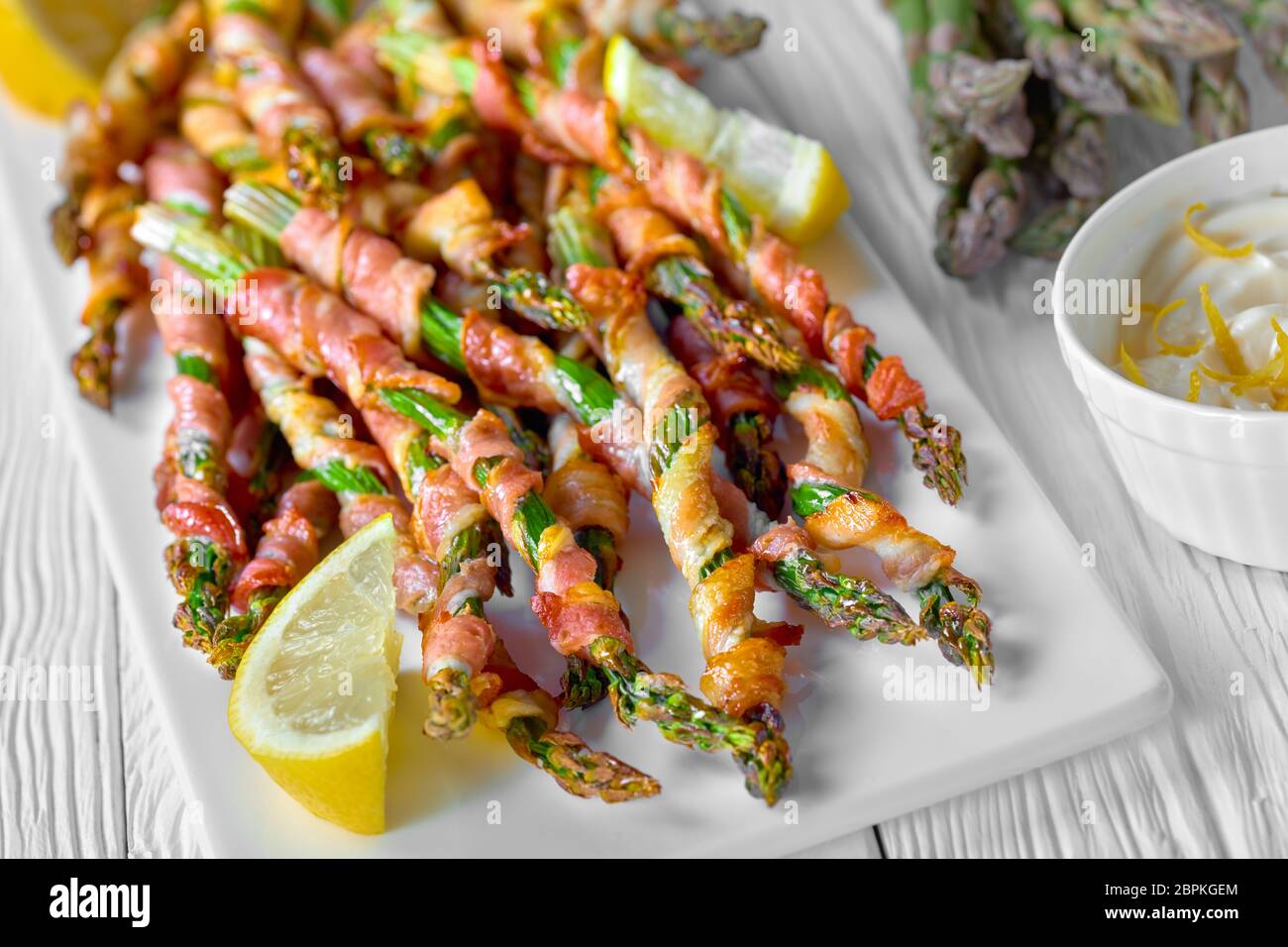 Garlic butter wrapped asparagus with bacon roasted with garlic and sea salt served on a white plate with lemon and sour cream sauce on a white wooden Stock Photo