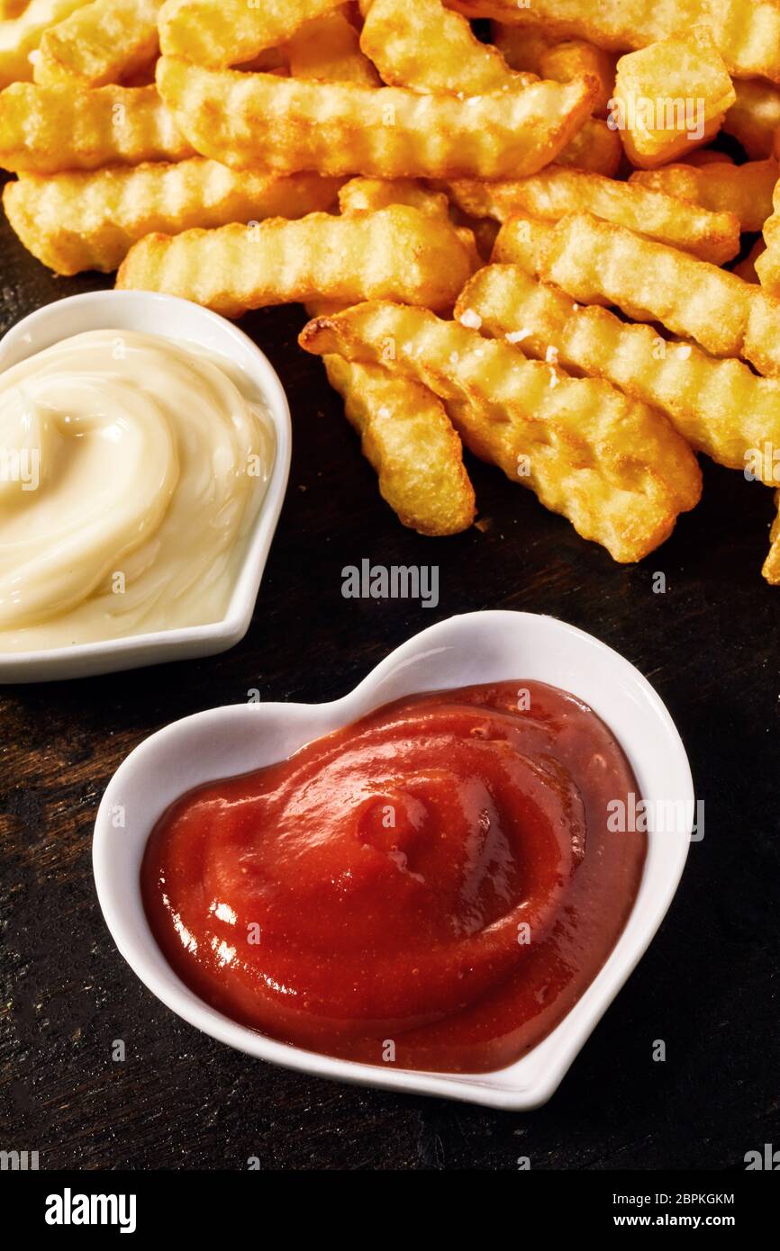 Heart-shaped bowls of ketchup, or tomato sauce, and creamy mayonnaise served as accompaniments to crispy golden crinkle cut potato chips or Pommes Fri Stock Photo