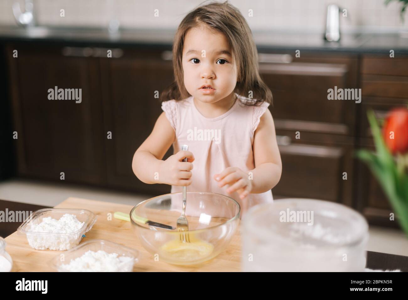 Happy cute little girl wearing white dress is whisking eggs in mixing