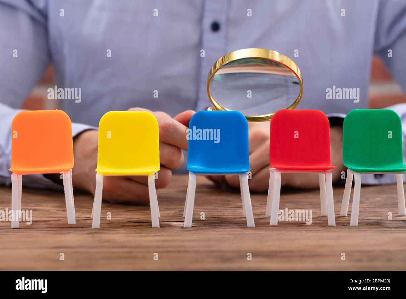 Close-up Of A Businessperson's Hand Looking At Vacant Colorful Chairs Through Magnifying Glass Stock Photo