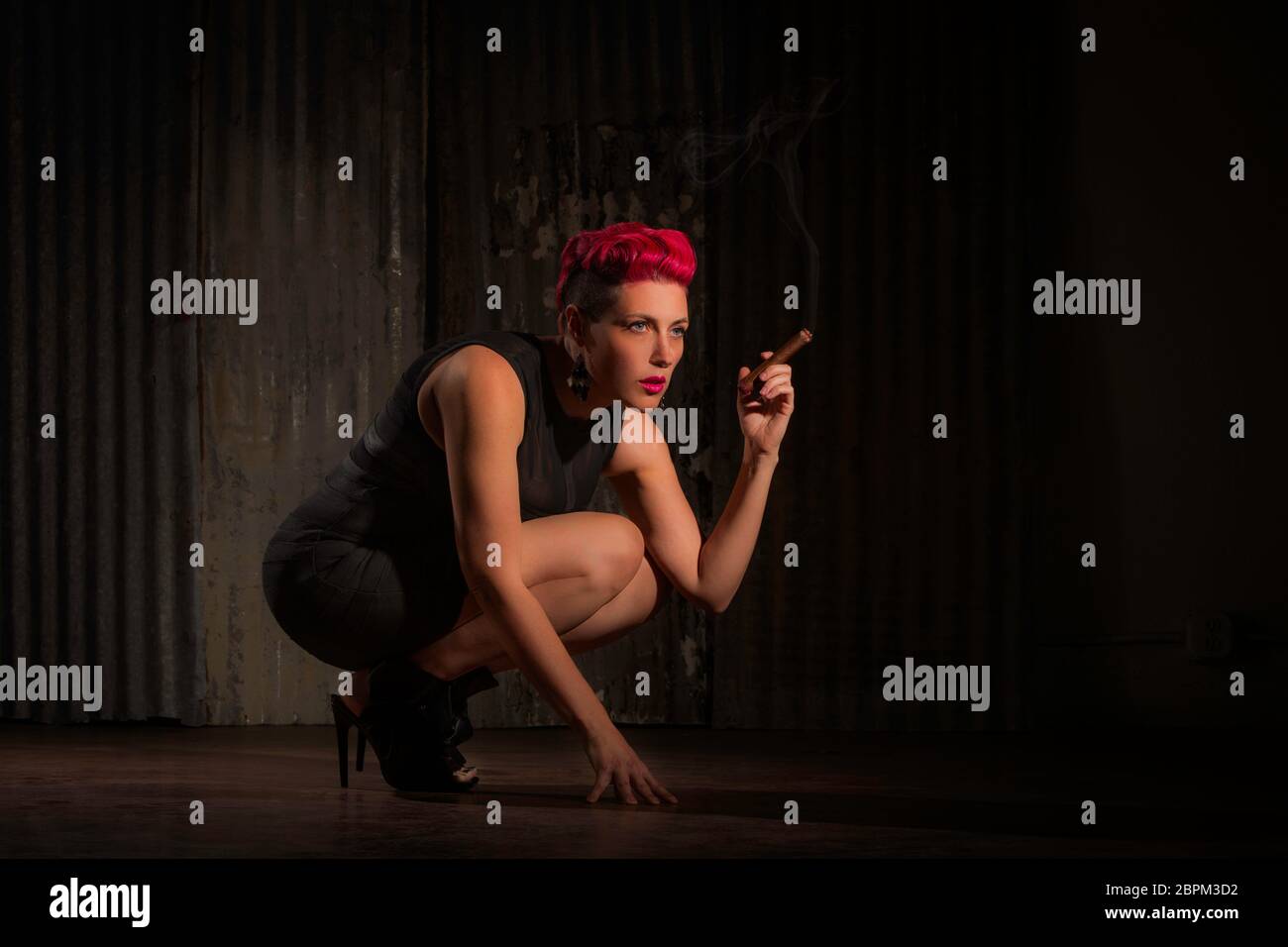 Sultry woman with fuschia hair crouching and holding a cigar Stock Photo