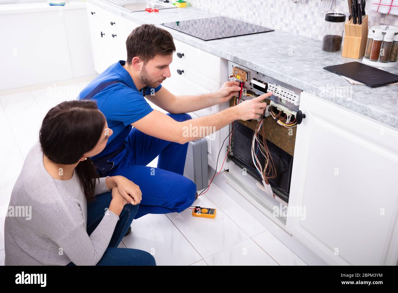 Woman Looking At Male Technician Checking Dishwasher With Digital Multimeter In Kitchen Stock Photo