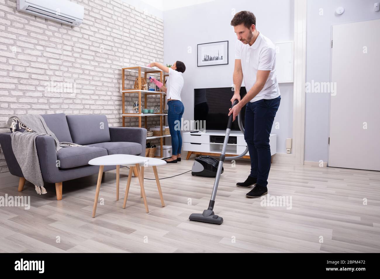 Side View Of Two Persons Cleaning The Shelf And Mopping Floor In The Living Room Stock Photo