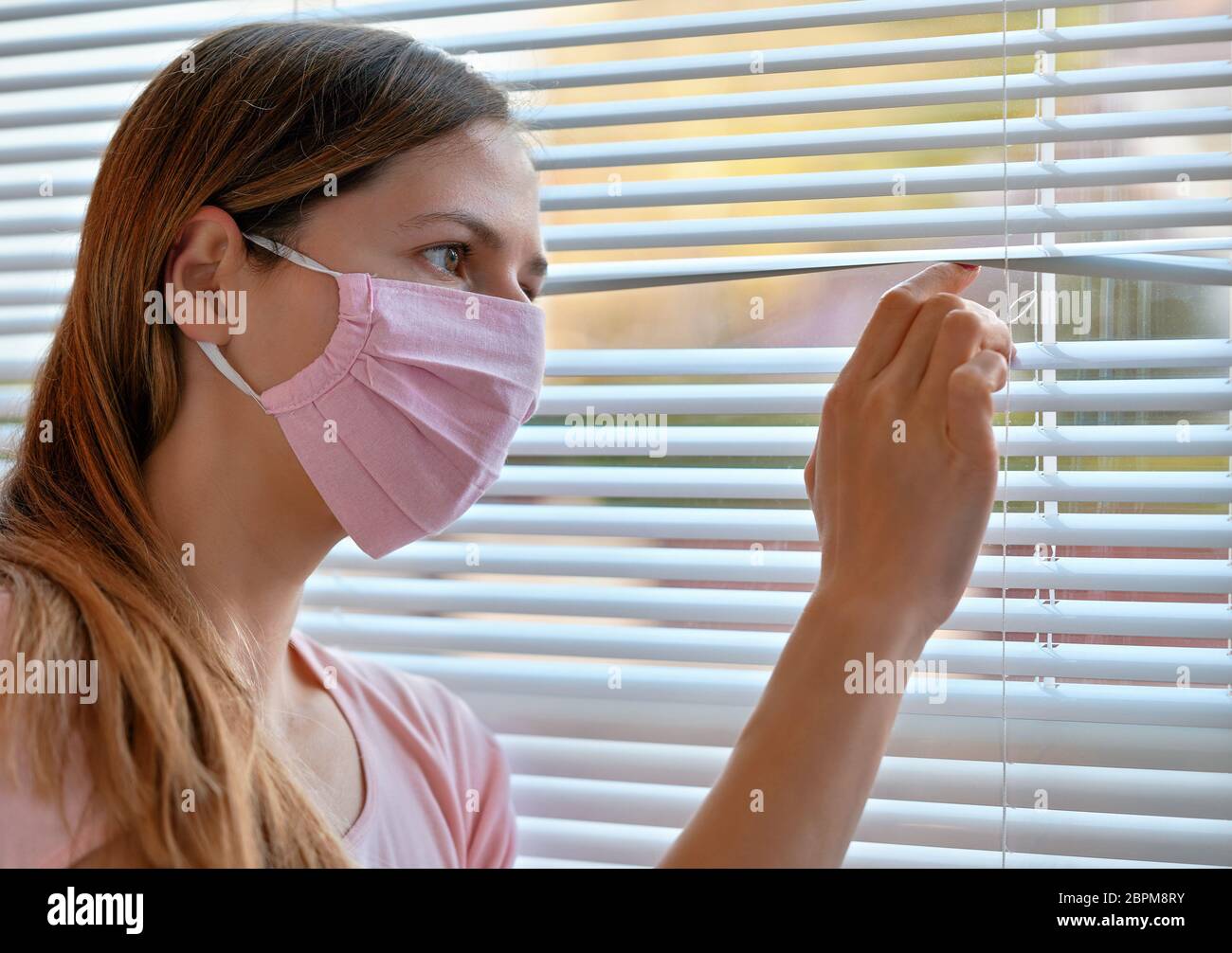 Young woman in pink home made cotton virus face mask, looking through window blinds outside. Quarantine or stay at home during coronavirus covid-19 ou Stock Photo