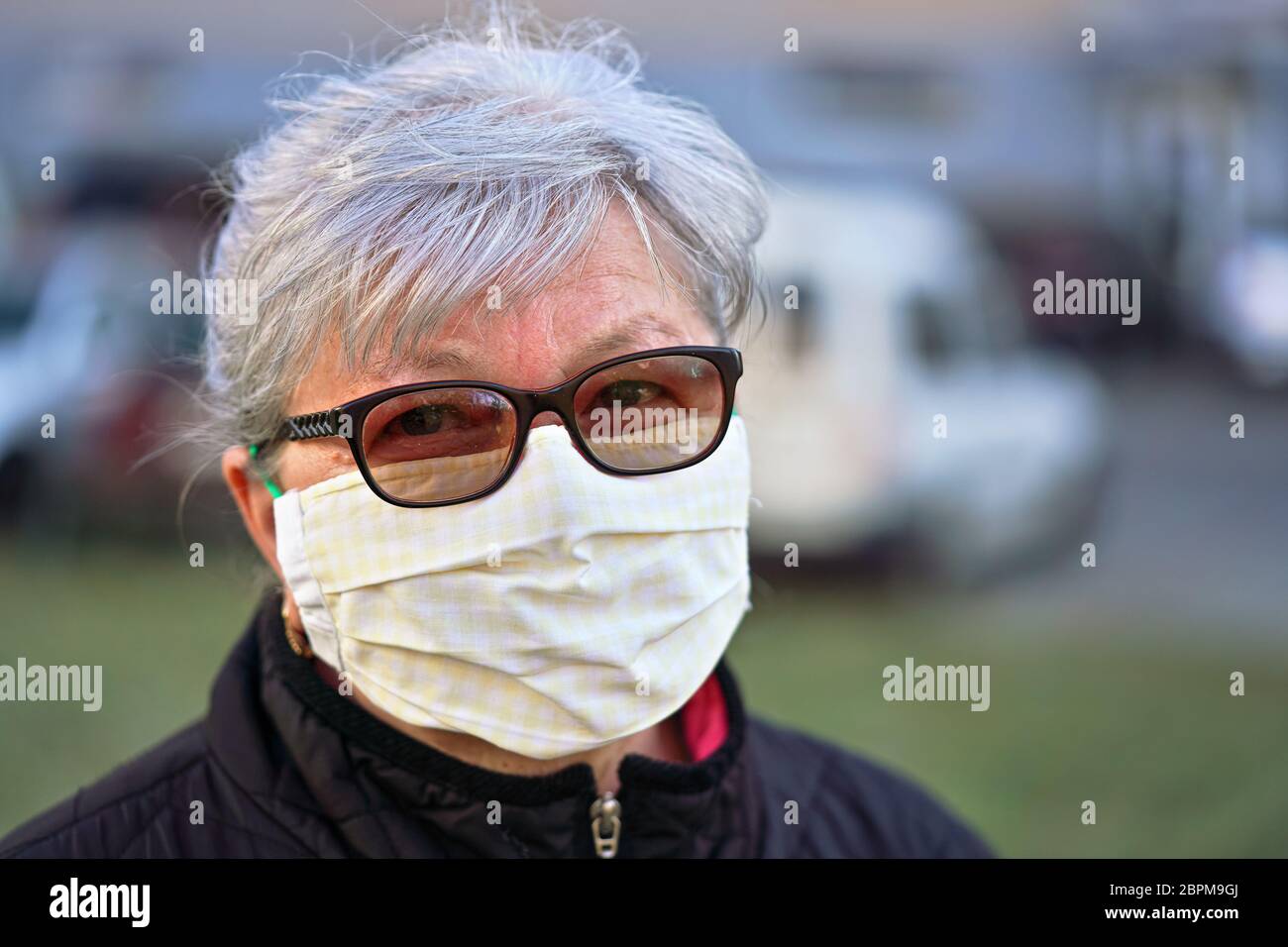 Elderly senior woman wearing home made cloth face mouth nose virus mask outside, blurred cars and building background. Can be used during coronavirus Stock Photo
