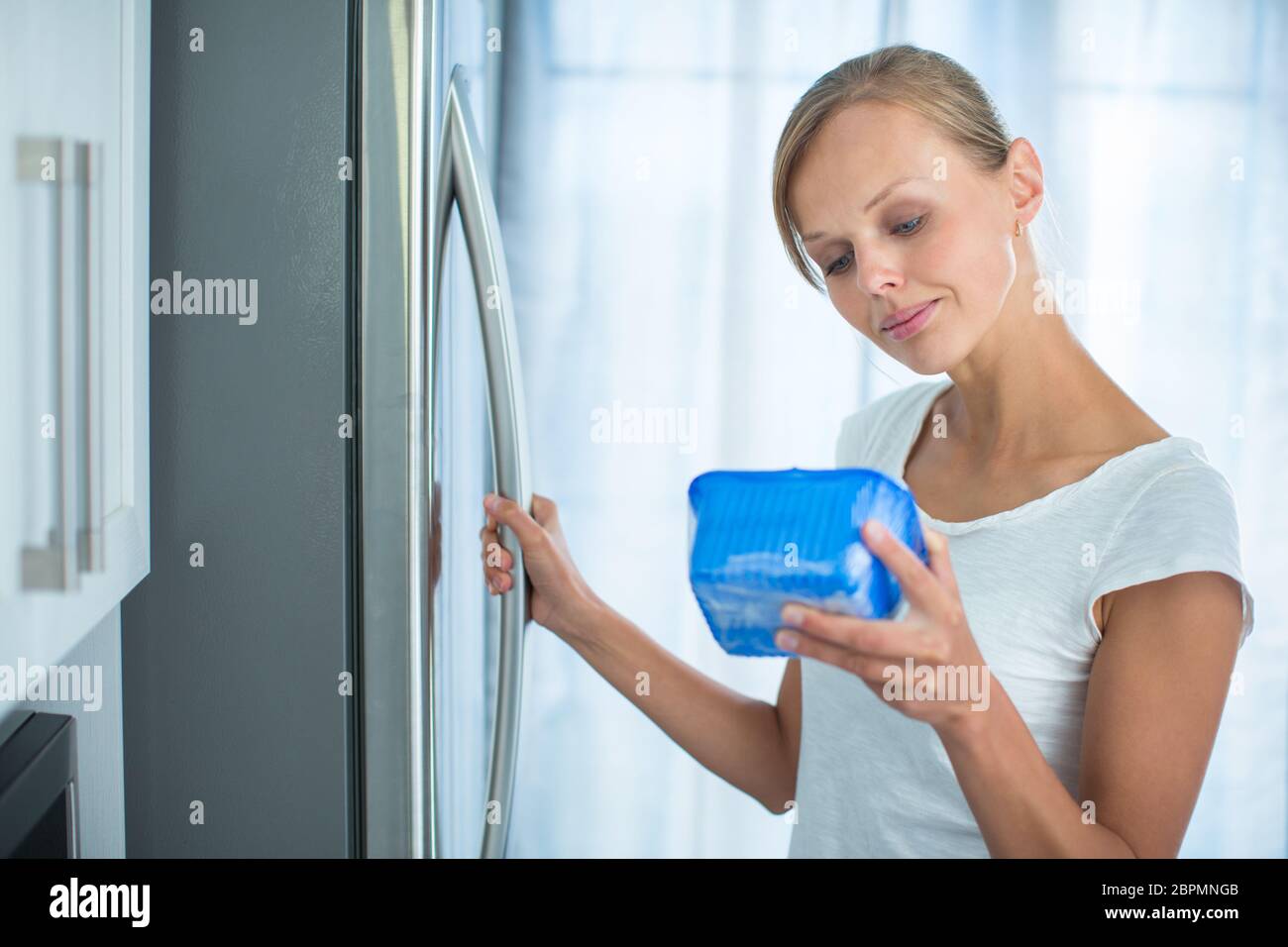 Is this still fine? Pretty, young woman in her kitchen by the fridge, looking at the expiry date of a product she took from her fridge - Stock Photo