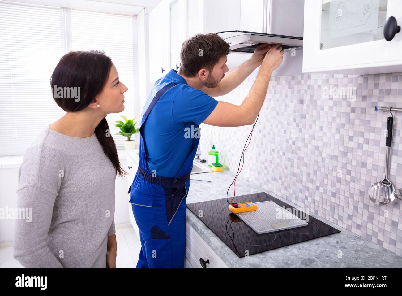 Male Repairman Checking Extractor Filter With Digital Multimeter While Woman Standing In Kitchen Stock Photo