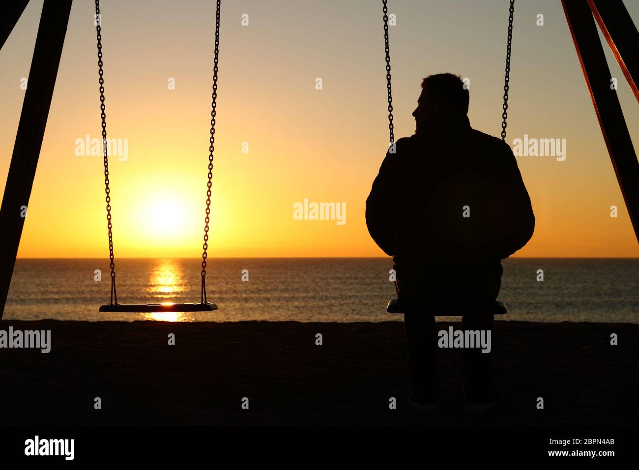 Back view backlighting silhouette of a man alone on a swing looking at empty seat at sunset on the beach in winter Stock Photo