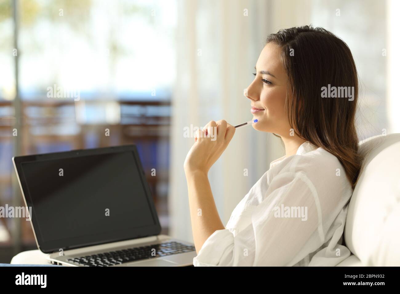 Side view portrait of a woman thinking looking at side and showing a blank laptop screen sitting on a couch of an apartment Stock Photo