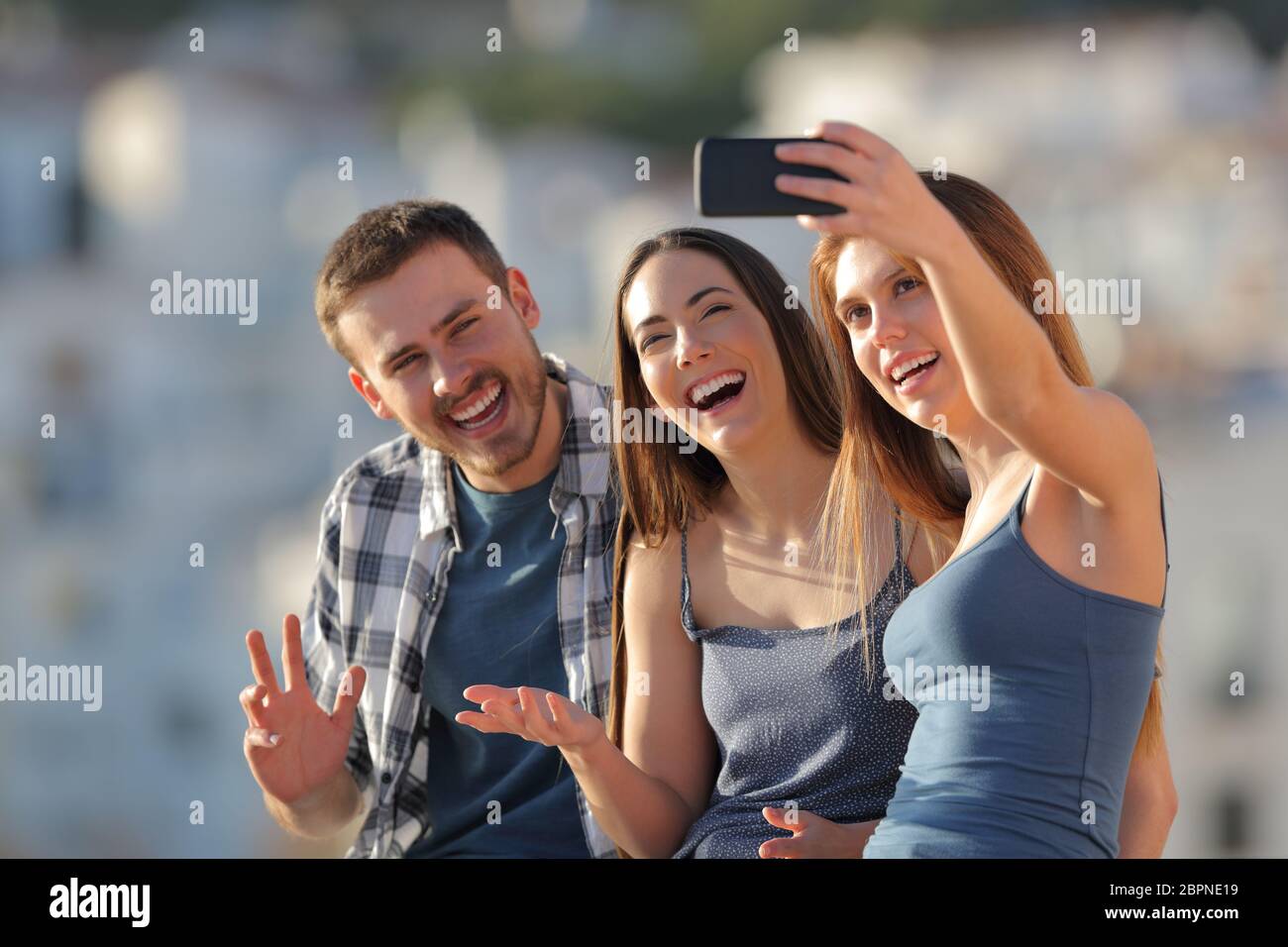 Three happy friends taking selfies with a smart phone outdoors in a town outskirts at sunset Stock Photo