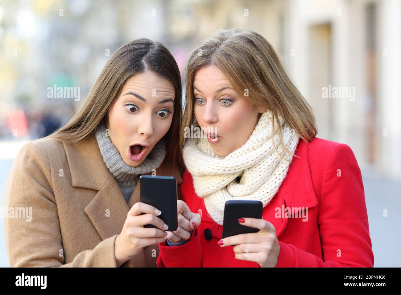 Front view portrait of two amazed women finding on line content on their smart phones on the street in winter Stock Photo