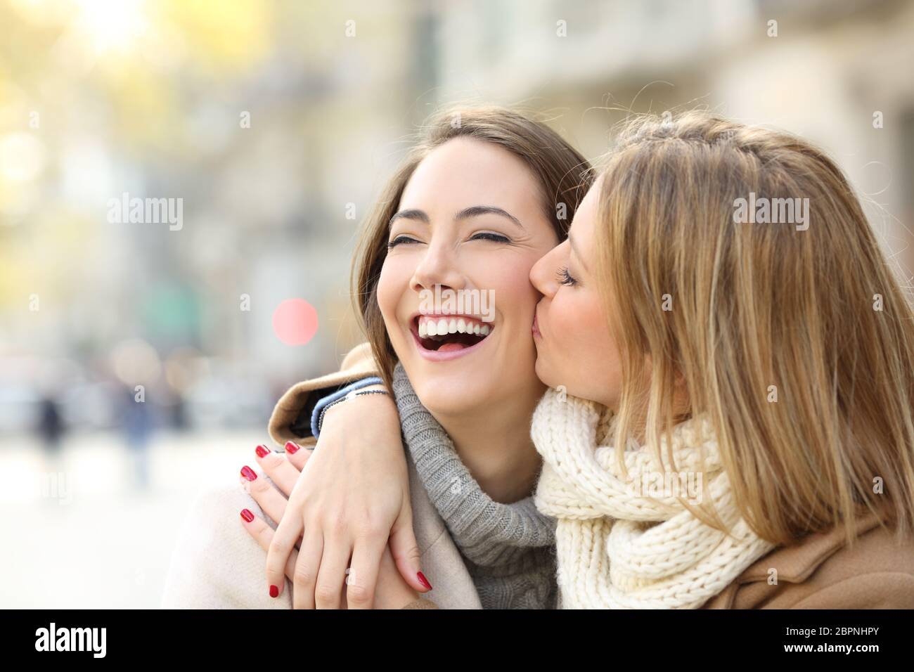 Affectionate girl kissing to her happy friend on the street in ...