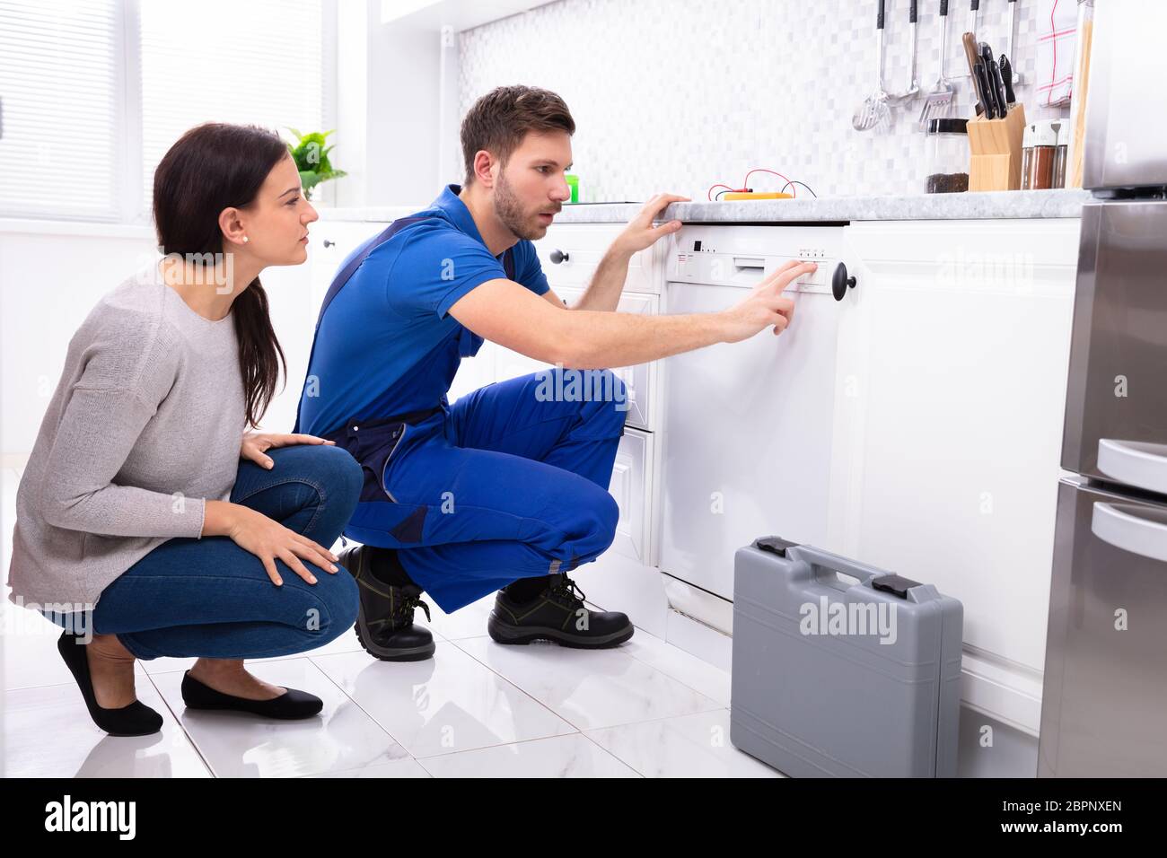 Serviceman Crouching On Kitchen Floor Pressing Button Of Dishwasher While Woman Looking At Him Stock Photo