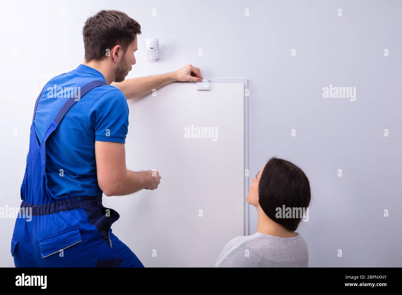 Happy Woman Looking At Electrician Installing Security System Door Sensor On Wall Stock Photo